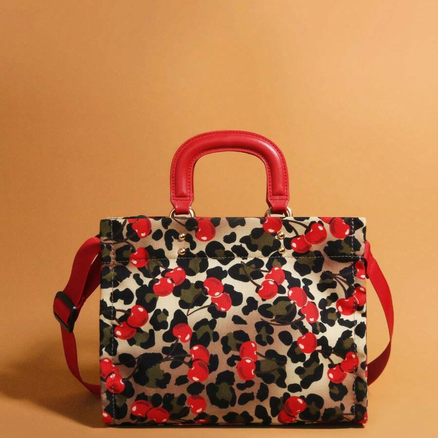 Front view of leopard-print handbag with red cherries, red handles and matching shoulder strap on ochre background