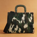 Front view of green and cream camouflage square handbag with olive handles and black strap on ochre background