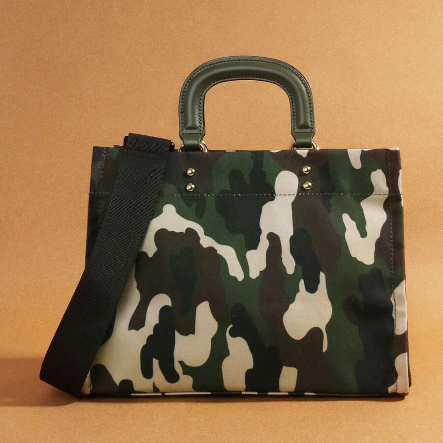 Front view of green and cream camouflage square handbag with olive handles and black strap on ochre background