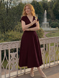 A woman stands on a balcony in a garden setting, wearing a sleeveless deep burgundy midi dress with a V-neckline and flared skirt. She is holding a glass of white wine and wearing cream-colored pointed-toe heels and gold jewelry.