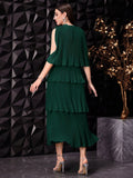 A woman modeling a dark green pleated tiered dress from the back, standing against a black geometric wall with a decorative gold plant on a marble pedestal.
