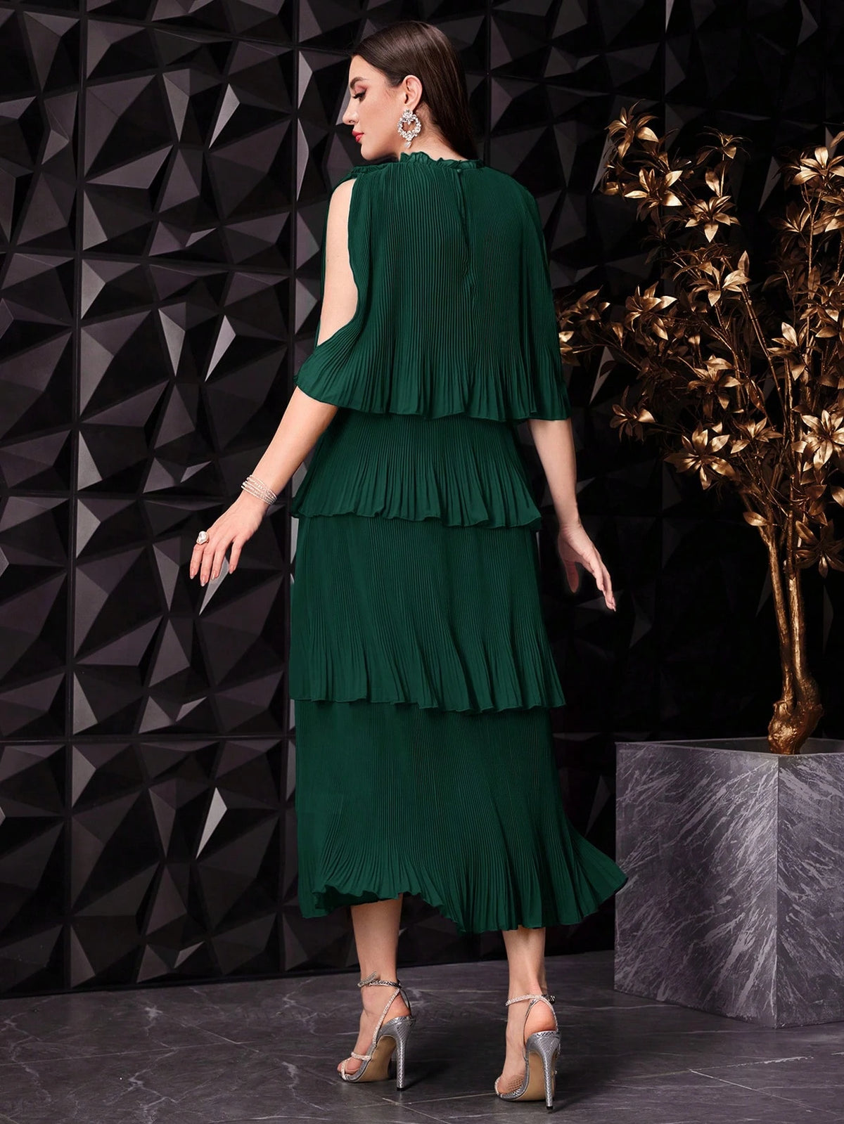 A woman modeling a dark green pleated tiered dress from the back, standing against a black geometric wall with a decorative gold plant on a marble pedestal.