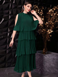 A woman models a dark green tiered pleated dress with a cold-shoulder sleeve, set against a dark geometric wall and a decorative gold tree.