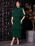 A woman models a dark green tiered dress with cold-shoulder sleeves and a pleated midi skirt, standing against a black geometric wall with a gold decorative tree on a marble pedestal.