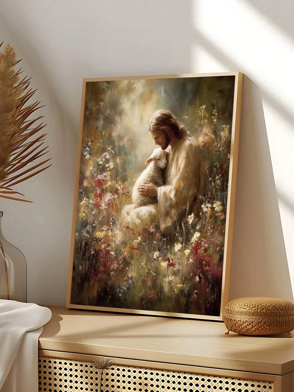 A framed vertical art print leaning against a wall on a wooden console table. The print depicts a serene, painterly scene of Jesus embracing a white sheep in a field of wildflowers.