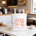 Cafe lifestyle view of beige bridal party canvas tote with pink orange beach graphic