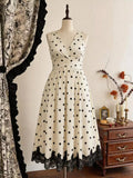 A cream-colored sleeveless A-line dress with black polka dots and a black lace hem, displayed on a mannequin in a vintage-style room next to a wooden chest and patterned curtains.