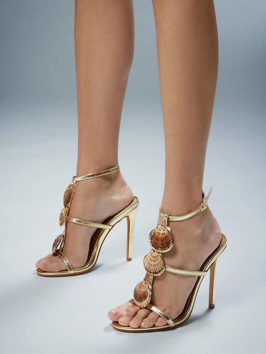 Front-side worn view of metallic gold scallop-shell T-strap square-toe stiletto sandals with ankle buckle for beach party styling