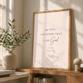 A framed wall art print with the text 'BE STILL, AND KNOW THAT I AM God. PSALM 46:10' in dark lettering on a warm off-white background, displayed in a light wood frame on a wooden console table in a sunlit room.