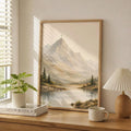 A framed landscape art print depicting a mountain peak with a cross, a calm lake, and pine trees, styled on a wooden dresser in a bright room next to a plant, books, a mug, and a lamp.