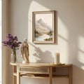 A framed landscape art print depicting a mountain and lake, styled on a light wood console table next to a purple flower vase, a religious statue, and a lit white candle in a sunlit room.