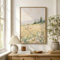 A framed landscape painting of a woman walking through a wildflower field hangs on a white wall above a wooden console table styled with a lamp, books, a candle, and a vase of white flowers.