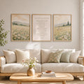 Three framed wall art prints hang above a light beige sofa in a bright, modern living room. The center print shows a Bible verse, while the side prints show soft landscape scenes with wildflowers and hills.