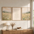Three framed wall art prints hang above a wooden console table in a softly lit room. The center print shows the Lord's Prayer, while the two side prints depict pastoral fields with mountains and a shepherd.