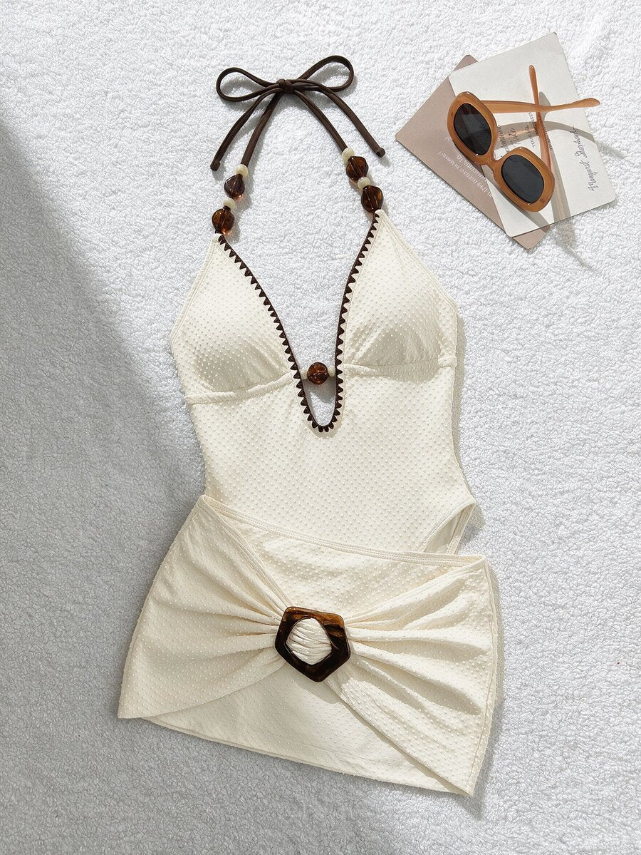 Flat lay of ivory halter swimsuit with scalloped brown trim and tortoiseshell-buckle draped skirt