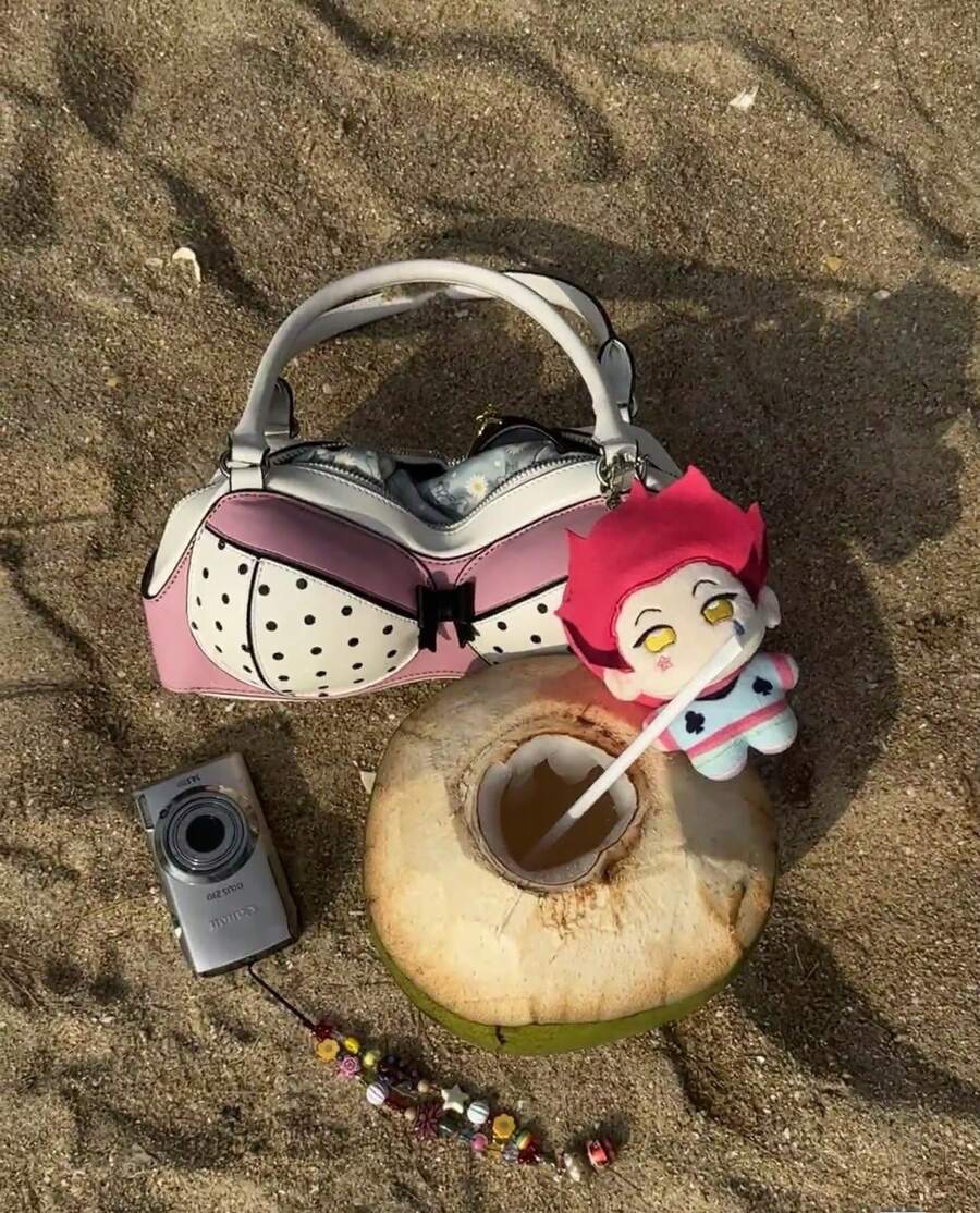 Pink polka-dot bra-shaped novelty handbag on beach sand beside a coconut drink, camera and plush charm