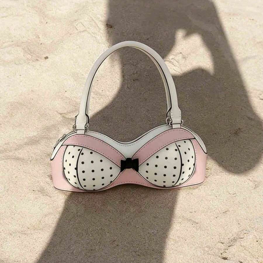 Full front view of pink bra-shaped handbag with black polka dots and white handle in sunlight
