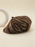 A brown and beige zebra-striped faux fur beret hat resting on a light beige surface next to a white ceramic ring prop.