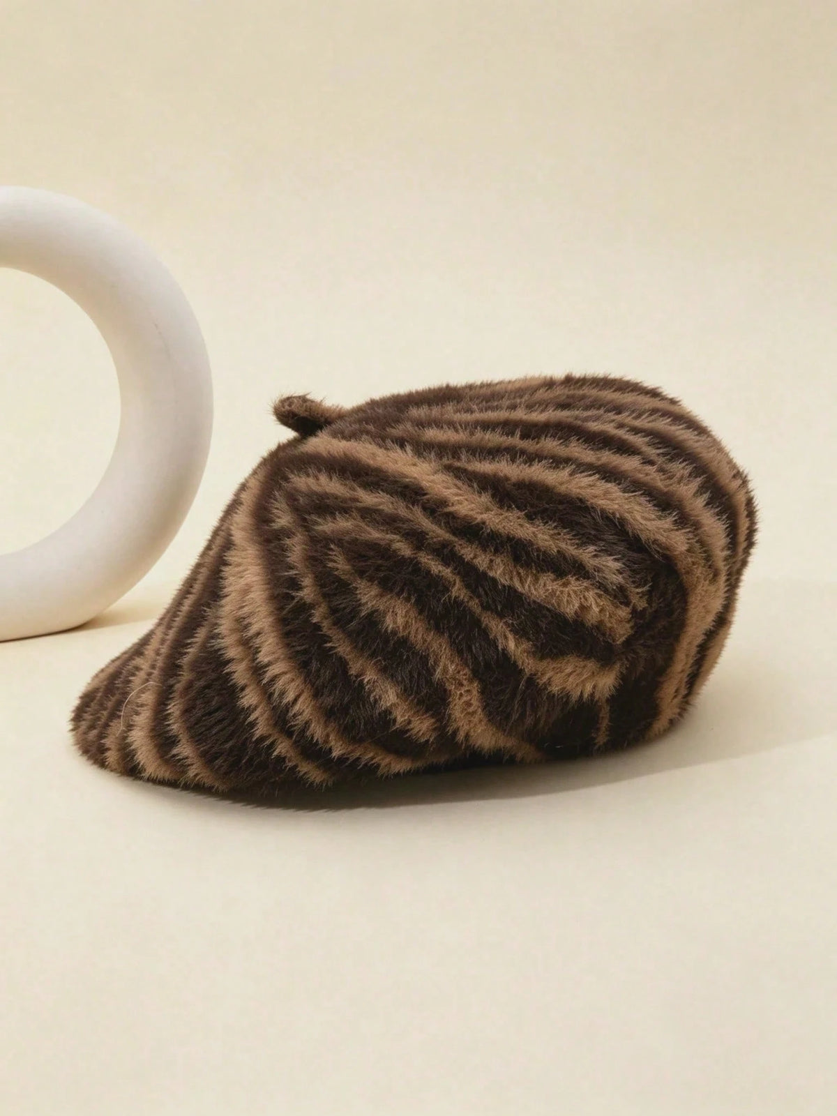 A brown and beige zebra-striped faux fur beret hat resting on a light beige surface next to a white ceramic ring prop.