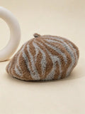 A fuzzy beret-style hat with brown and light gray zebra-like stripes, photographed on a warm beige background with a partial white sculptural prop at the left edge.