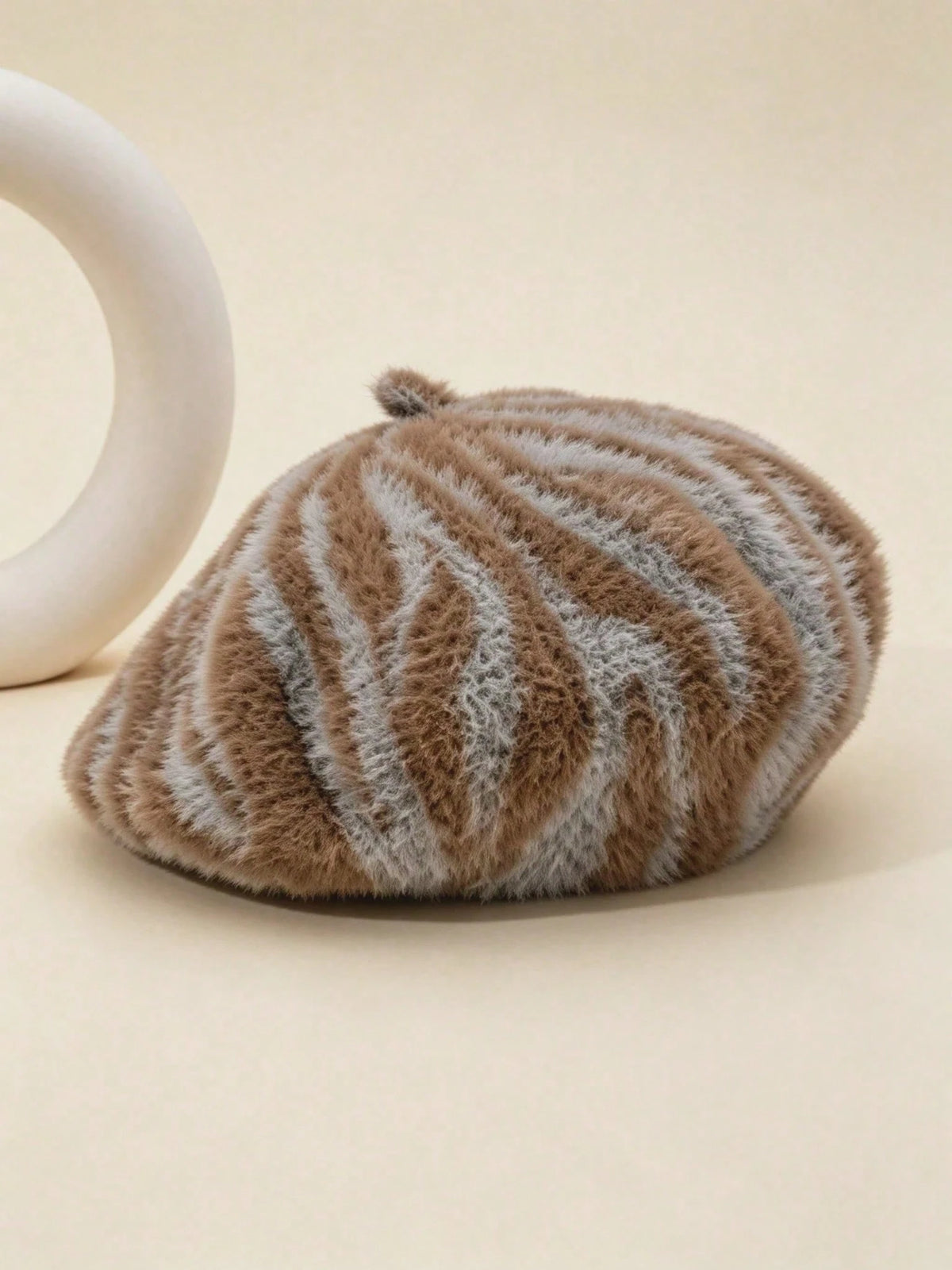 A fuzzy beret-style hat with brown and light gray zebra-like stripes, photographed on a warm beige background with a partial white sculptural prop at the left edge.