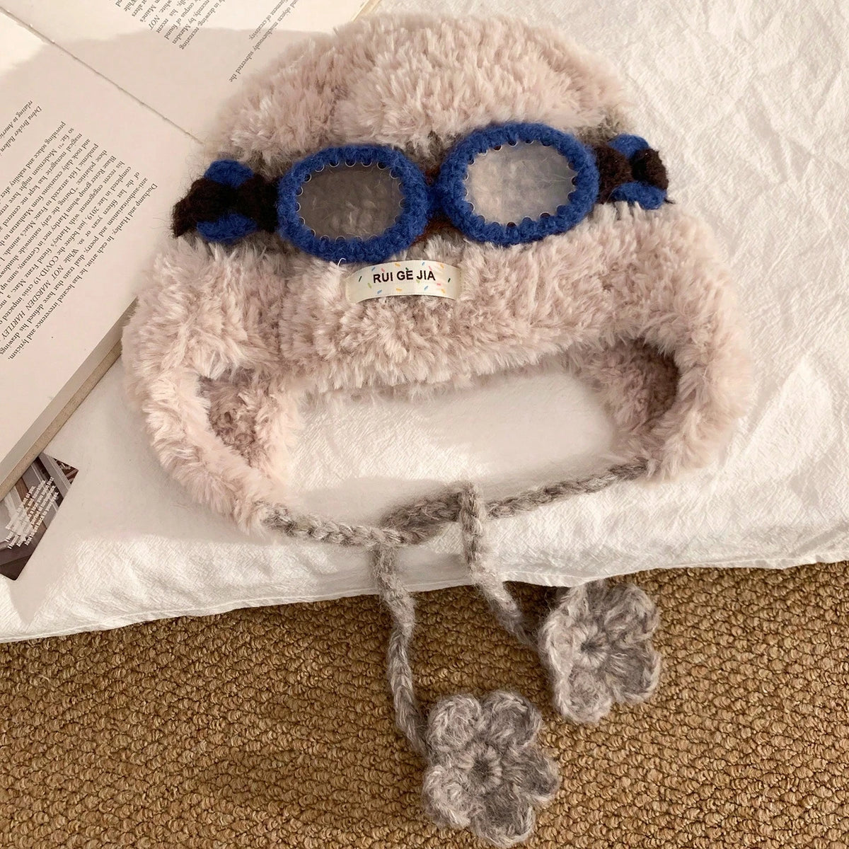 Top view of cream fuzzy trapper hat with blue goggles and long braided ties ending in knit flowers