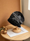 A black and gray marled knit beanie hat with a ribbed brim, displayed on a wooden stand on a light wood table next to dried roses.