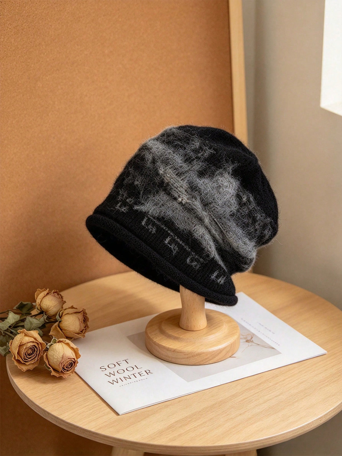 A black and gray marled knit beanie hat with a ribbed brim, displayed on a wooden stand on a light wood table next to dried roses.