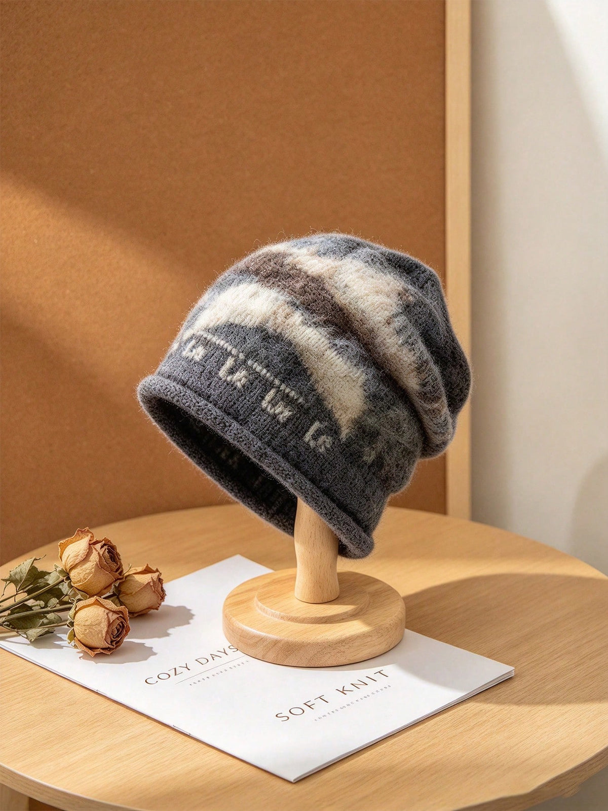 A slouchy knit beanie hat in gray, cream, and brown tones with a knitted pattern, displayed on a wooden stand on a light wood table.