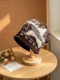 A fuzzy knit beanie hat in brown and cream tones with a knitted pattern, displayed on a wooden stand on a round wooden table.