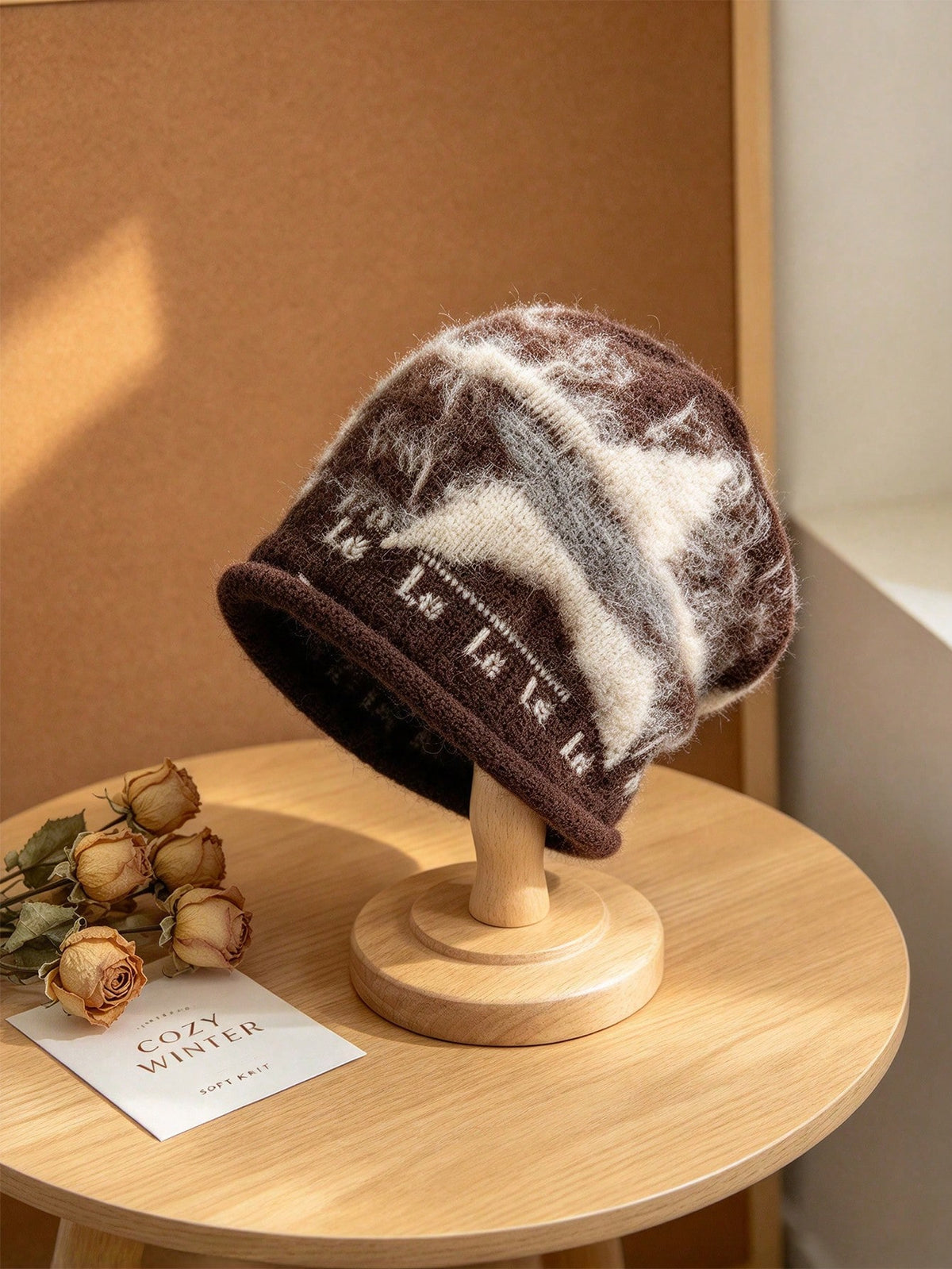 A fuzzy knit beanie hat in brown and cream tones with a knitted pattern, displayed on a wooden stand on a round wooden table.