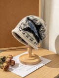 A knit beanie hat displayed on a wooden stand on a tabletop. The hat is cream with black and blue marled patterning and black script text reading 'De La La La La'.