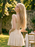 A woman with long blonde hair stands outdoors in a garden wearing a short cream-colored dress with ruffled straps and a lace-up back. The background features green foliage, a stone wall, and a dining table with chairs.