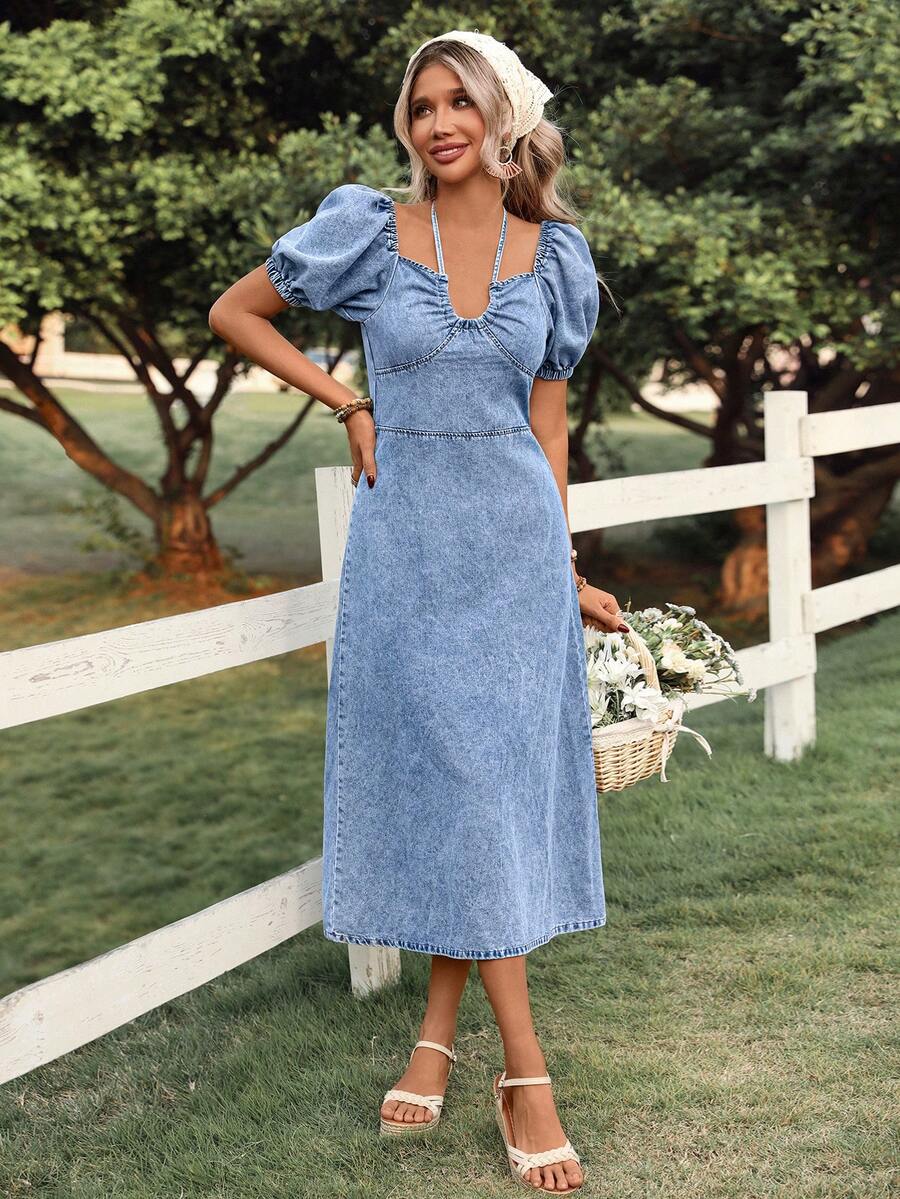 Front three-quarter view of blue acid-wash denim midi dress with puff sleeves and fitted waist in a garden
