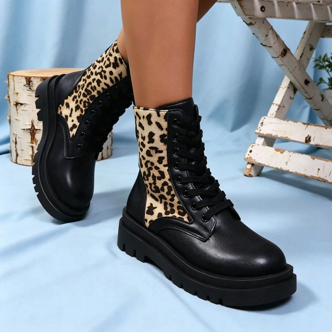 A pair of black lace-up ankle boots with leopard-print fabric panels on the sides, shown worn on a model's feet against a light blue backdrop with rustic props.