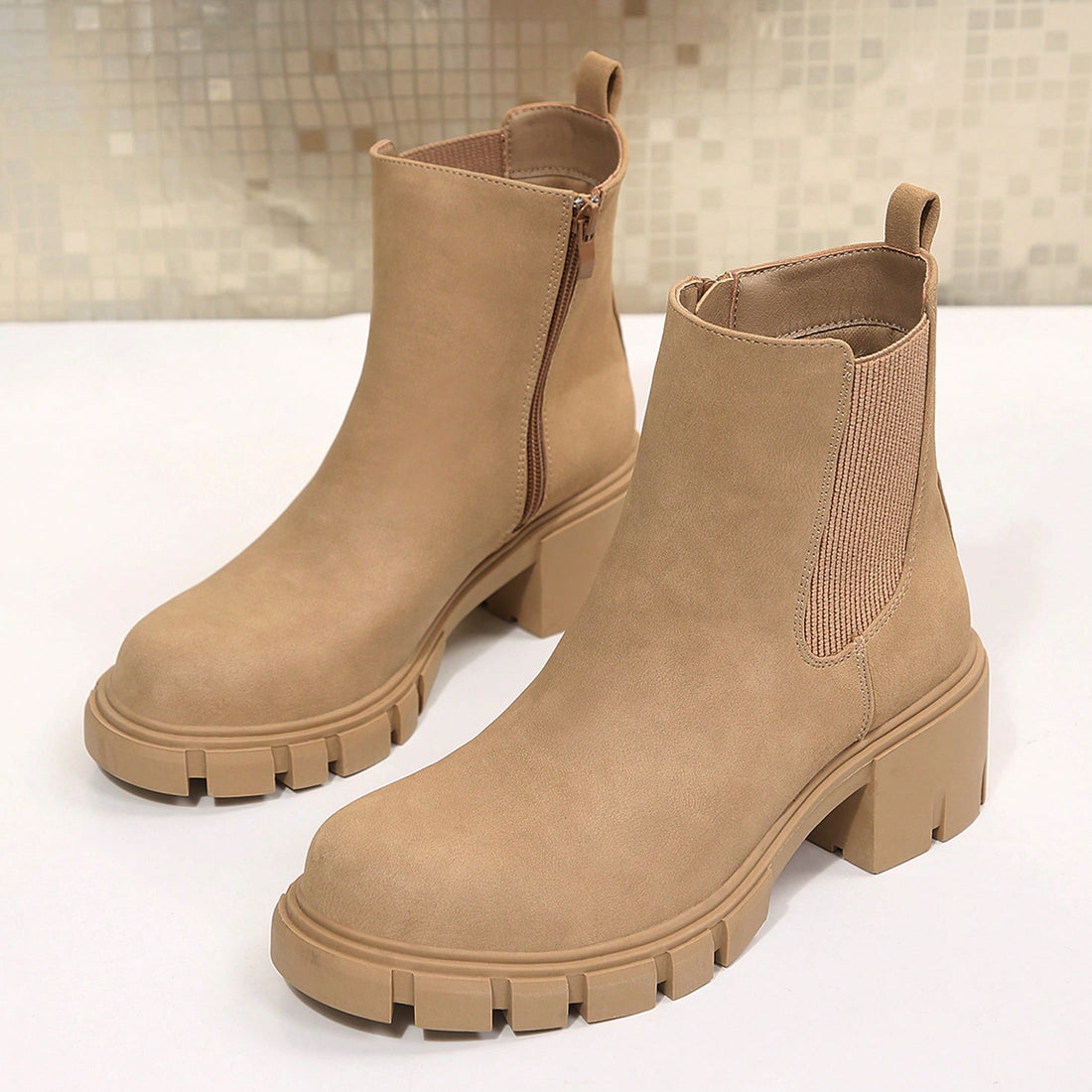 A pair of beige faux-suede ankle boots with chunky lug soles and elastic side panels, displayed on a white surface with a tiled background.