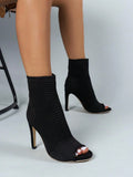 Black knit ankle boots with open toes and high stiletto heels, shown on a model's feet against a light gray studio background.