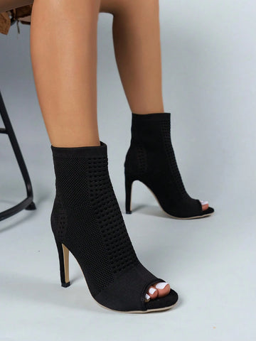 Black knit ankle boots with open toes and high stiletto heels, shown on a model's feet against a light gray studio background.