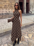 Full view of the brown plaid sleeveless halter dress with long A-line skirt, styled with boots outside a stone building.