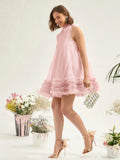 A woman models a light pink sleeveless tulle dress with a ruffled hem, paired with metallic gold high-heeled sandals and a small silver clutch, set against a clean white background with decorative floral arrangements.