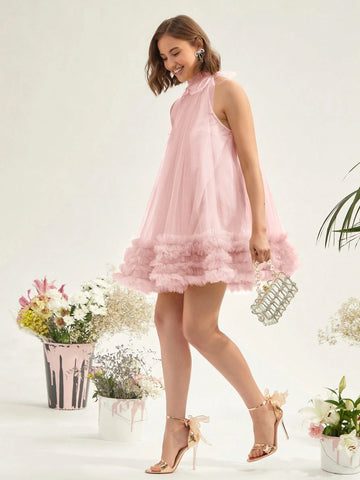 A woman models a light pink sleeveless tulle dress with a ruffled hem, paired with metallic gold high-heeled sandals and a small silver clutch, set against a clean white background with decorative floral arrangements.