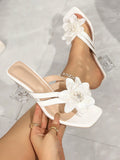 White strappy mule sandals with large fabric flowers and clear block heels, shown being worn and held against a light gray tiled floor.
