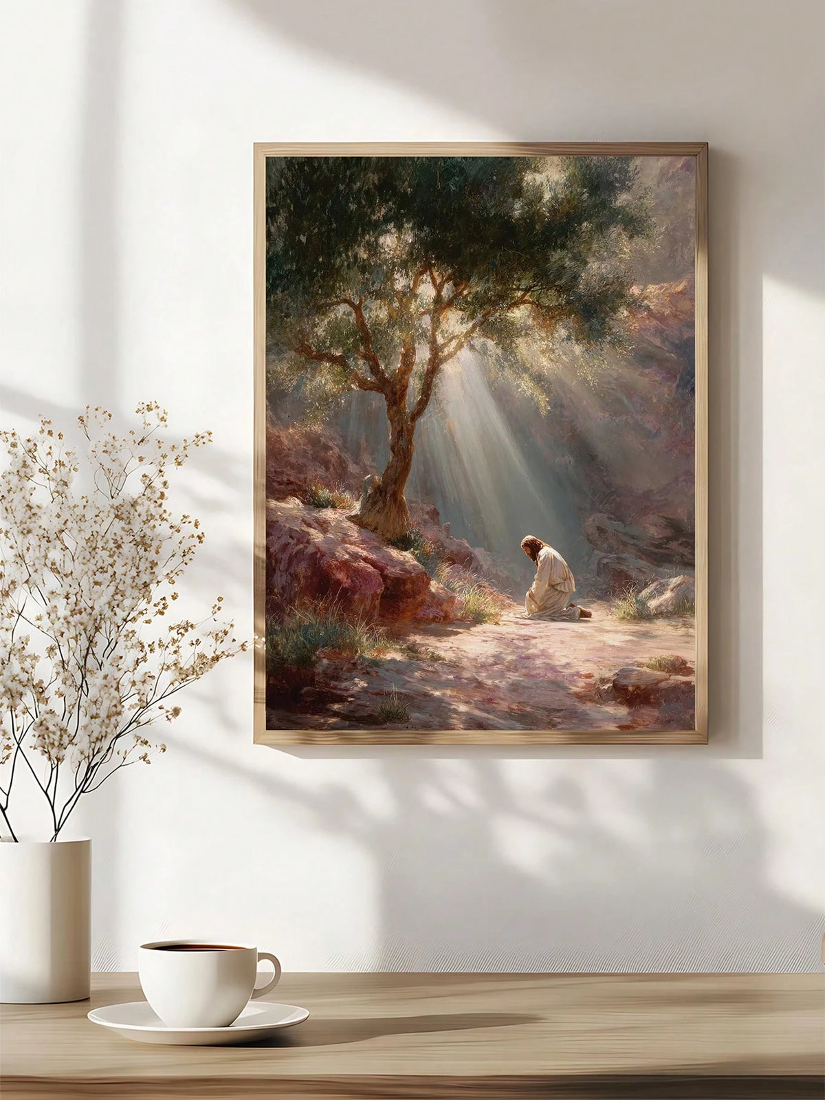 A framed art print depicting a serene outdoor landscape with sunbeams filtering through trees, hanging on a white wall above a wooden table with a cup and vase of dried flowers.