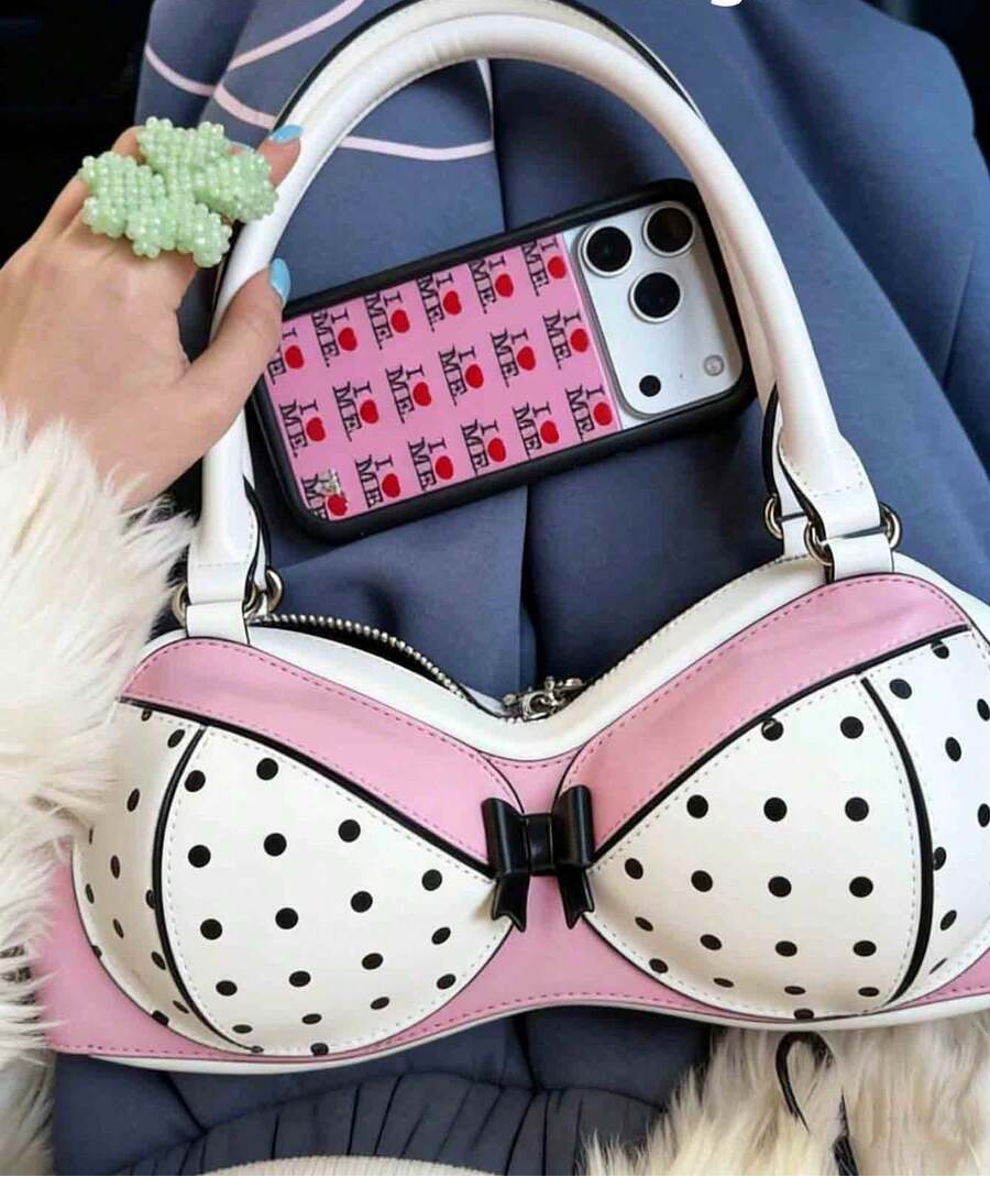 Close-up of pink and white bra-shaped handbag with black polka dots, bow and white handles beside a phone