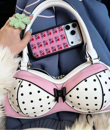 Close-up of pink and white bra-shaped handbag with black polka dots, bow and white handles beside a phone