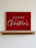 Red canvas wall art reading “MERRY Christmas” in white serif and cursive lettering, displayed in a light wood frame above a console shelf