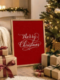 Styled front view of red Merry Christmas calligraphy canvas in a light wood frame beside a Christmas tree and wrapped gifts