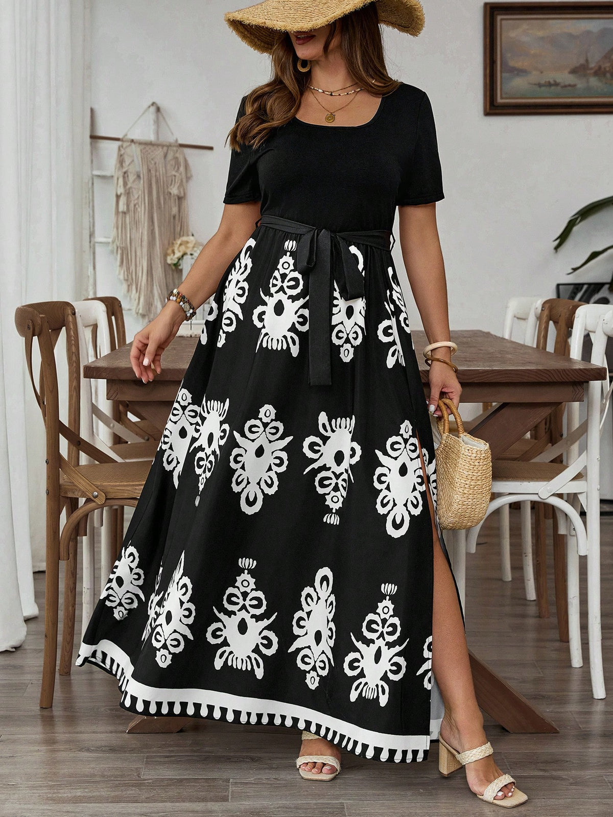 A woman models a black short-sleeve maxi dress with a white damask-style floral pattern and a tassel hem, styled with a straw hat, beige sandals, and a woven handbag in a dining room setting.