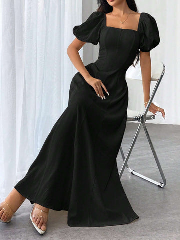 A woman wearing a black maxi dress with puff sleeves and a square neckline, posing next to a metal folding chair in front of white curtains.