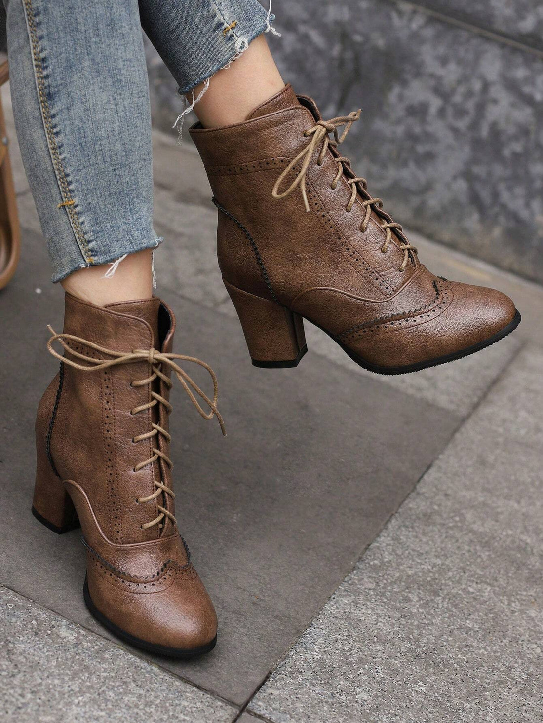 A pair of brown lace-up ankle boots with block heels and brogue detailing, shown worn with frayed blue jeans on a gray stone pavement.
